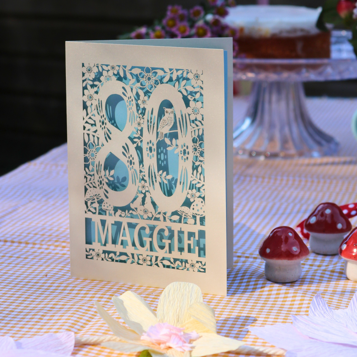 A handmade 80th birthday card with intricate laser-cut floral patterns and the number 80, standing on a checkered tablecloth with decorative paper flowers and mushroom figurines.