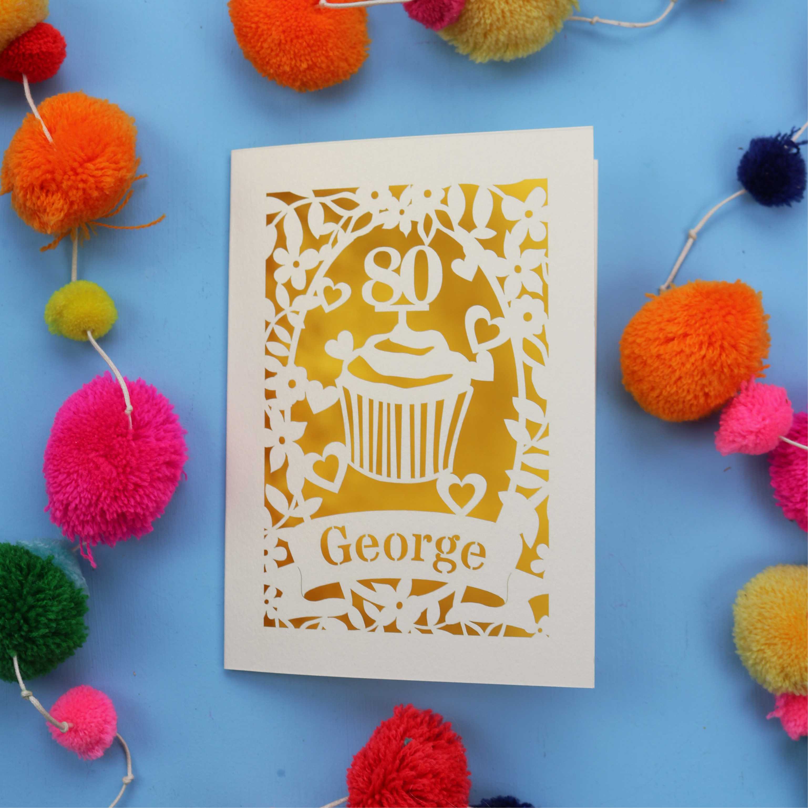 Laser cut 80th Birthday Card with '80' cut out cupcake design on a blue background with colourful pom-poms.
