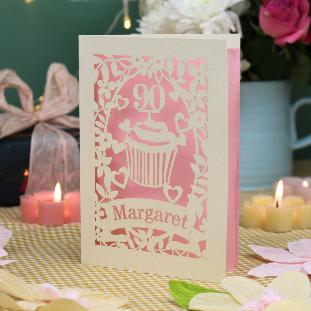 A personalized laser-cut birthday card with a pink cupcake design and the name 'Margaret' on a 90th birthday, displayed on a yellow-and-white checked tablecloth with lit candles and flowers in the background.