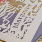 Close-up of a cream personalized laser-cut paper card featuring the name 'Margaret' in a banner with floral and heart motifs, layered over a lavender and gold background.