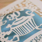 A close-up of a laser-cut paper cupcake design with a blue background and cream cut out details, including the number 90 and decorative hearts.