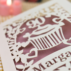 A close-up of a cream laser-cut cupcake design with floral and heart cut outs, set against a dusky pink background with a softly blurred pink candle in the upper left.