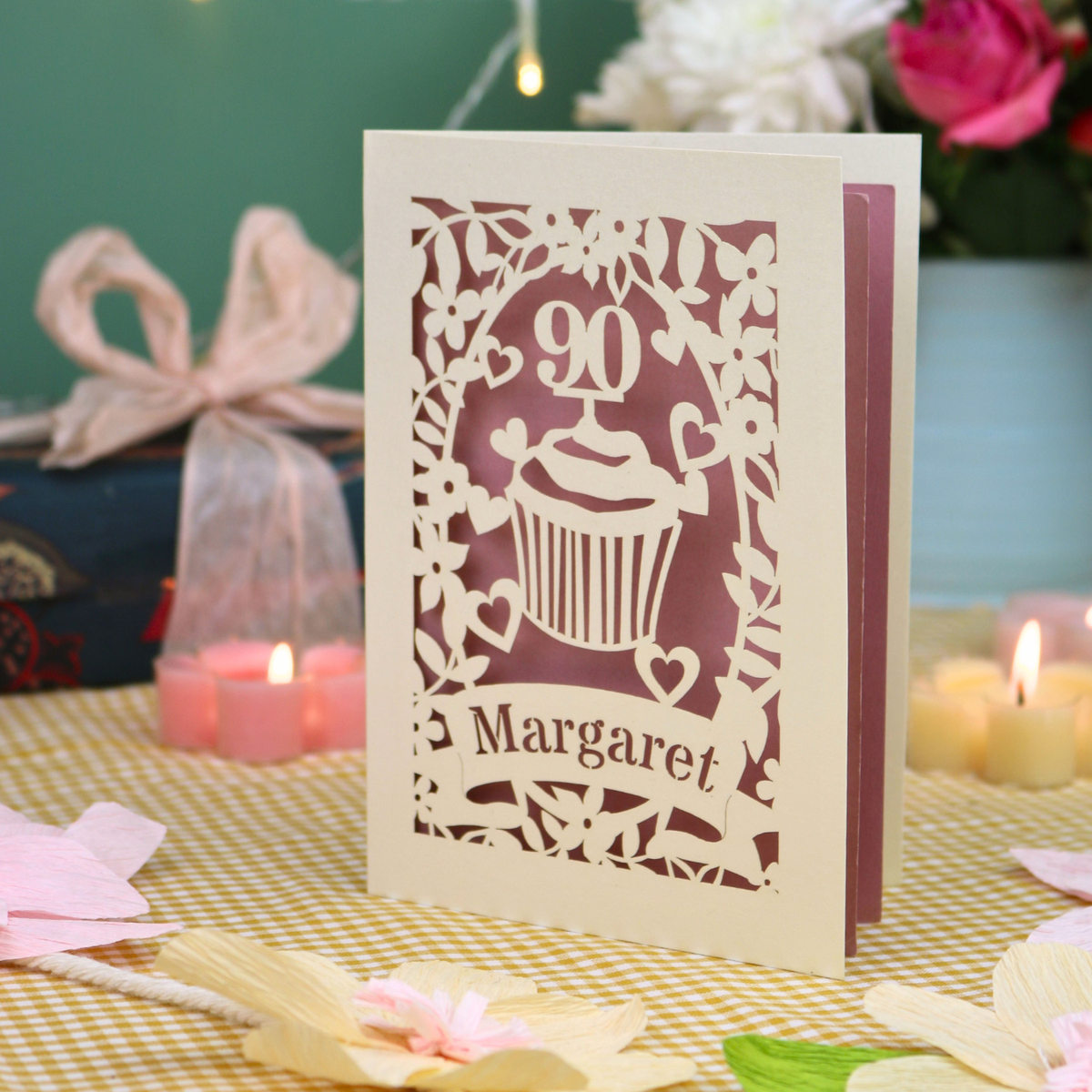 A personalized laser-cut 90th birthday  card featuring a cupcake design with the number 90 and the name 'Margaret', displayed on a checked tablecloth with candles and flowers in the background.