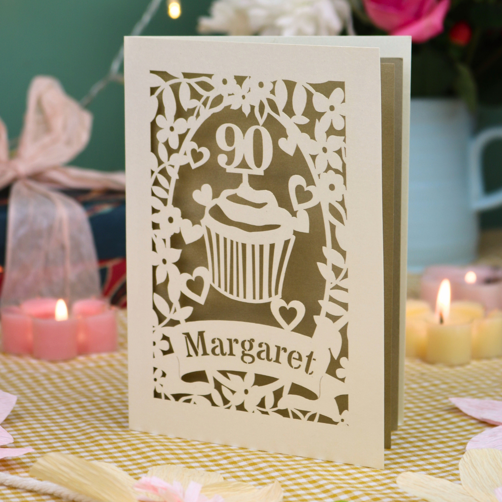 A personalised cut out 90th birthday  card featuring a cupcake design with the number 90 and the name 'Margaret', displayed on a checked tablecloth with candles and flowers in the background.