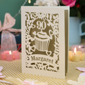 A personalised cut out 90th birthday  card featuring a cupcake design with the number 90 and the name 'Margaret', displayed on a checked tablecloth with candles and flowers in the background.