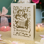 A personalised cut out 90th birthday  card featuring a cupcake design with the number 90 and the name 'Margaret', displayed on a checked tablecloth with candles and flowers in the background.