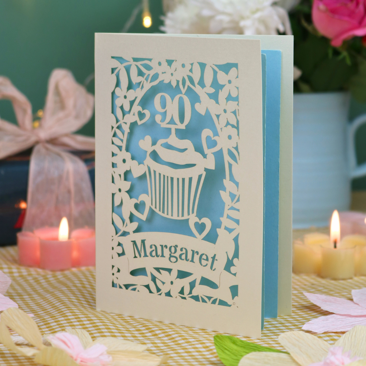 A personalised laser-cut birthday card for Margaret, celebrating a 90th birthday with a cupcake design, displayed on a checked tablecloth with lit candles and flowers in the background.