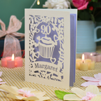 A laser-cut birthday card for a 90th birthday, featuring a cupcake and floral design with the name 'Margaret' on a checked tablecloth with candles in the background.