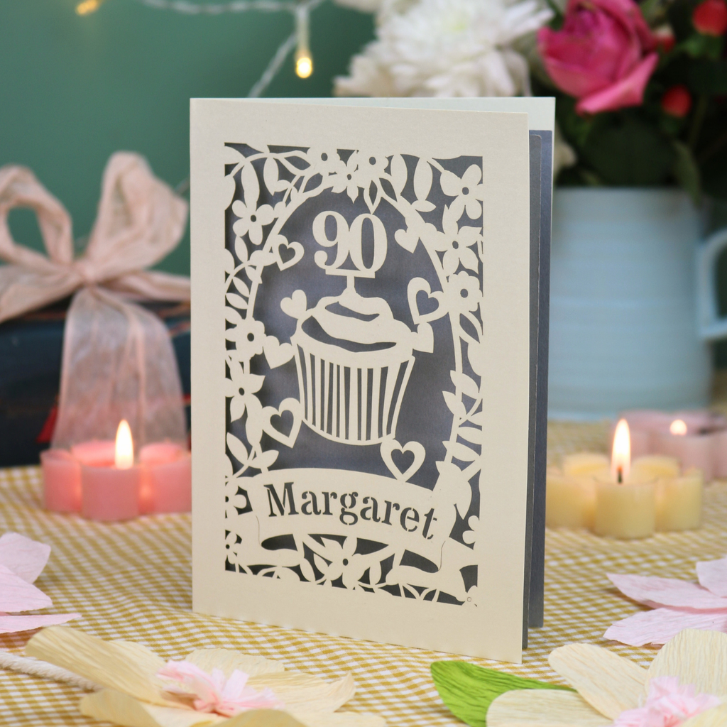 A personalised 90th birthday card featuring a cupcake design with the number 90 and the name 'Margaret', displayed on a checked tablecloth with candles and flowers in the background.