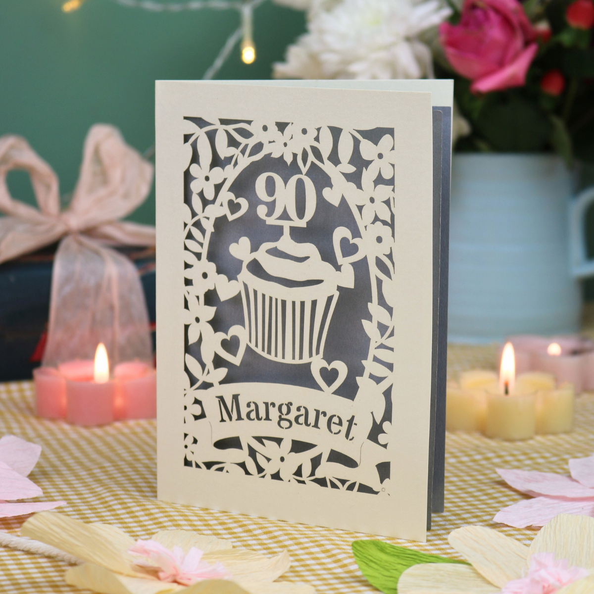 A personalised 90th birthday card featuring a cupcake design with the number 90 and the name 'Margaret', displayed on a checked tablecloth with candles and flowers in the background.