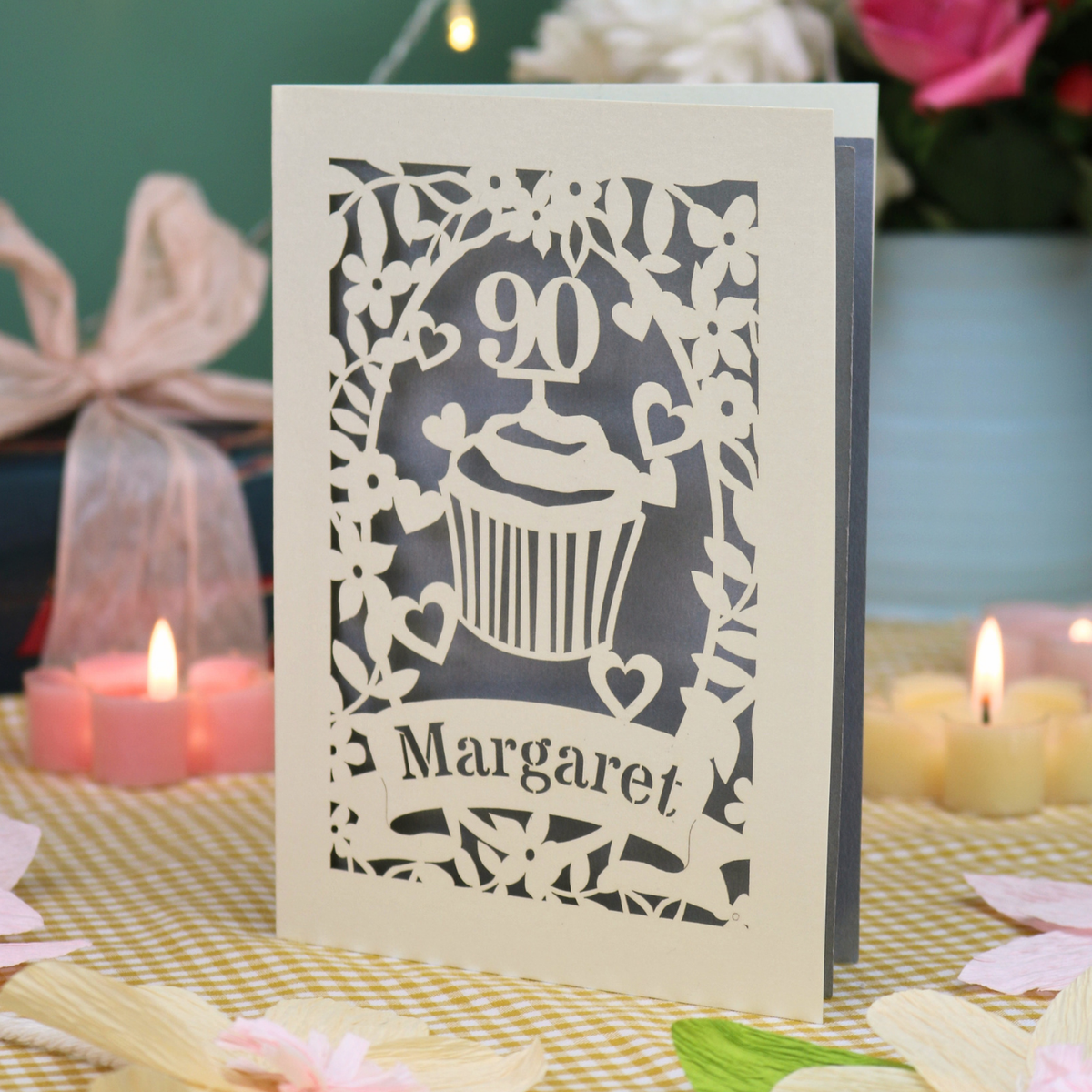 A personalized laser-cut birthday card featuring a cupcake and floral design with the name 'Margaret' and the number '90', displayed on a yellow-and-white checked tablecloth with lit candles in the background.