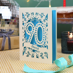 A personalized 90th birthday card with intricate laser-cut cream floral and number '90' designs, featuring the name 'David' on a banner. The card is shown standing on a checked tablecloth with a turquoise ribbon, a lit candle, and a disco ball in the background.