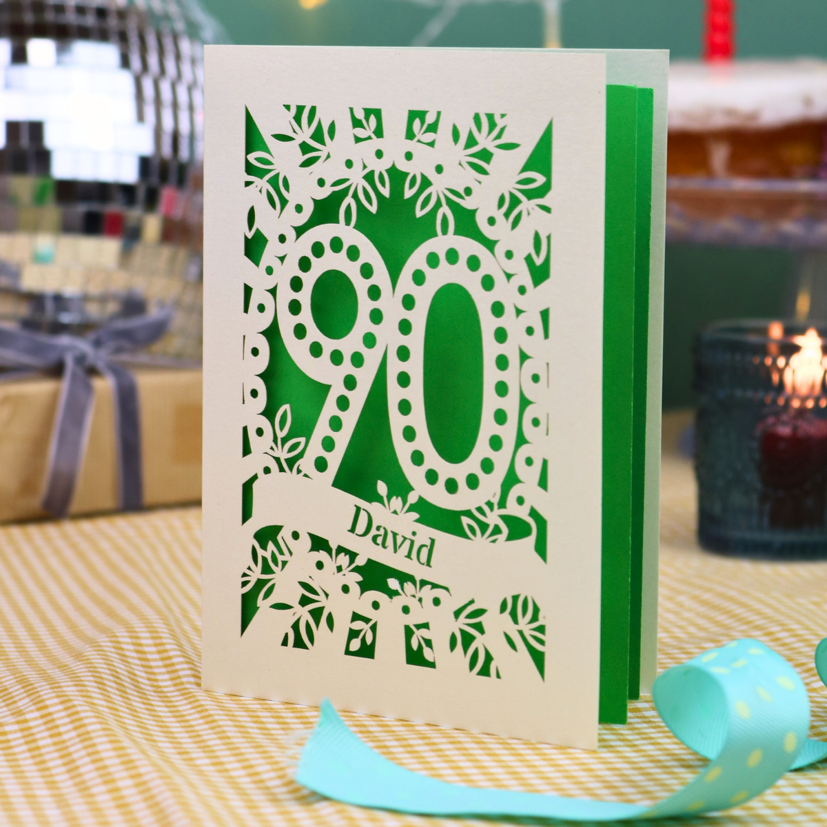 A personalised keepsake 90th birthday card with intricate laser-cut cream floral and number '90' designs, featuring the name 'David' on a banner. The card is shown standing on a checked tablecloth with a turquoise ribbon, a lit candle, and a disco ball in the background.
