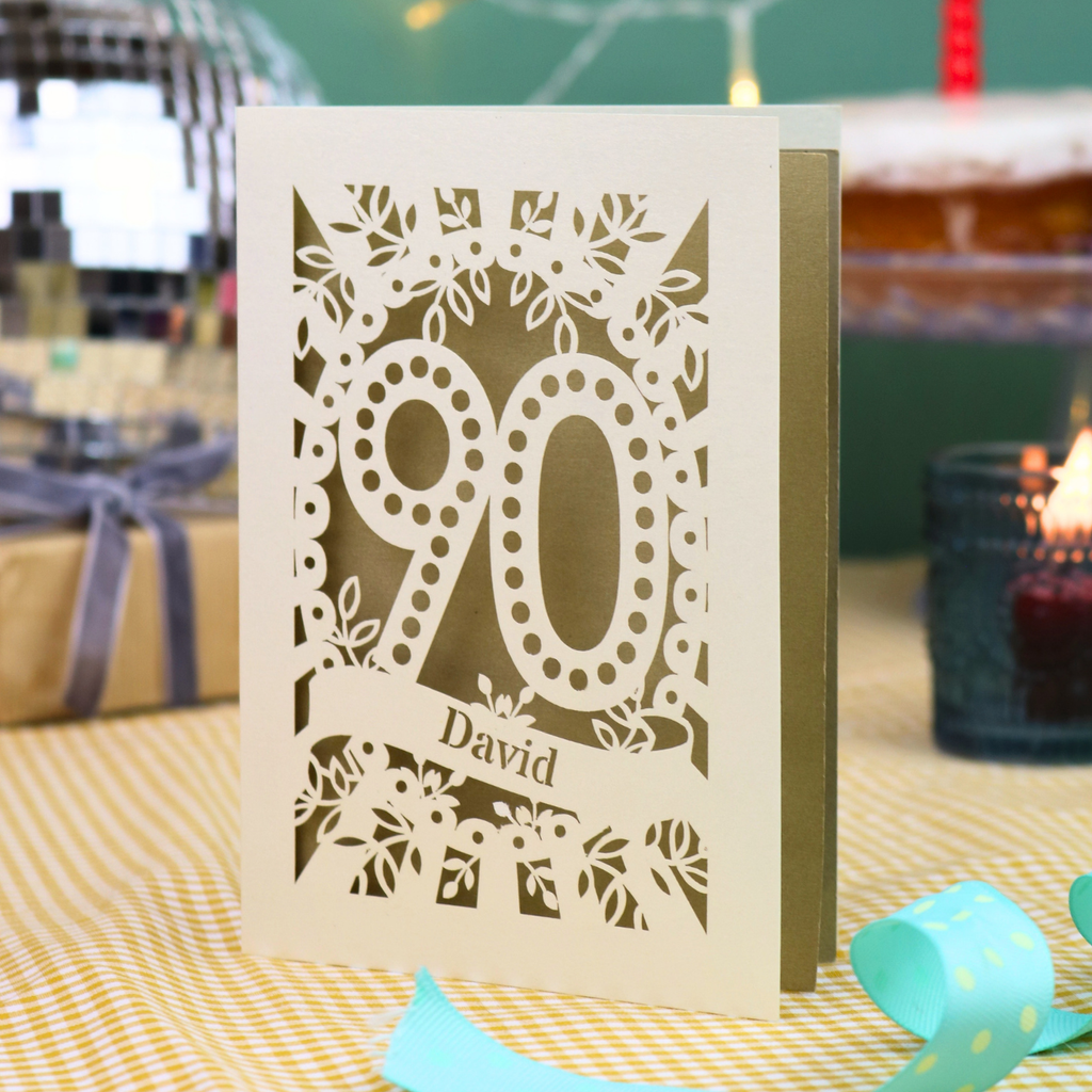 A personalized 90th birthday card with intricate laser-cut cream floral and number '90' designs, featuring the name 'David' on a banner. The card is shown standing on a checked tablecloth with a turquoise ribbon, a lit candle, and a disco ball in the background.