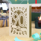 A personalized 90th birthday card with intricate laser-cut cream floral and number '90' designs, featuring the name 'David' on a banner. The card is shown standing on a checked tablecloth with a turquoise ribbon, a lit candle, and a disco ball in the background.