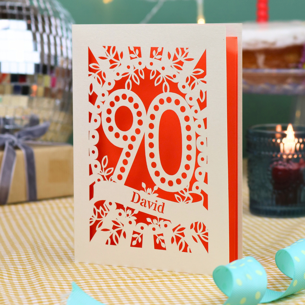 A personalized 90th birthday card with intricate laser-cut cream floral and number '90' designs, featuring the name 'David' on a banner. The card is shown standing on a checked tablecloth with a turquoise ribbon, a lit candle, and a disco ball in the background.