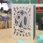 A personalized 90th birthday card with intricate laser-cut cream floral and number '90' designs, featuring the name 'David' on a banner. The card is shown standing on a checked tablecloth with a turquoise ribbon, a lit candle, and a disco ball in the background.