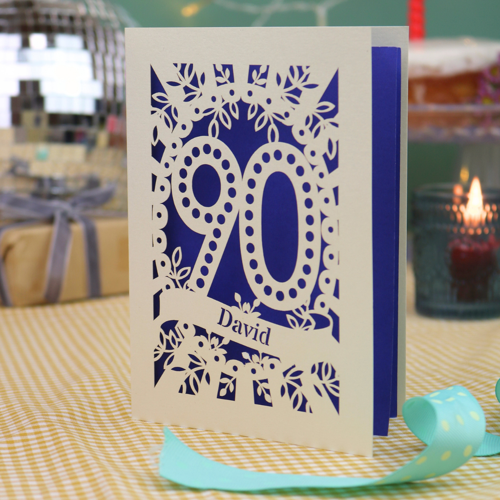 A personalized 90th birthday card with intricate laser-cut cream floral and number '90' designs, featuring the name 'David' on a banner. The card is shown standing on a checked tablecloth with a turquoise ribbon, a lit candle, and a disco ball in the background.