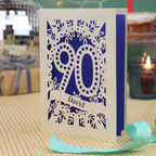 A personalized 90th birthday card with intricate laser-cut cream floral and number '90' designs, featuring the name 'David' on a banner. The card is shown standing on a checked tablecloth with a turquoise ribbon, a lit candle, and a disco ball in the background.