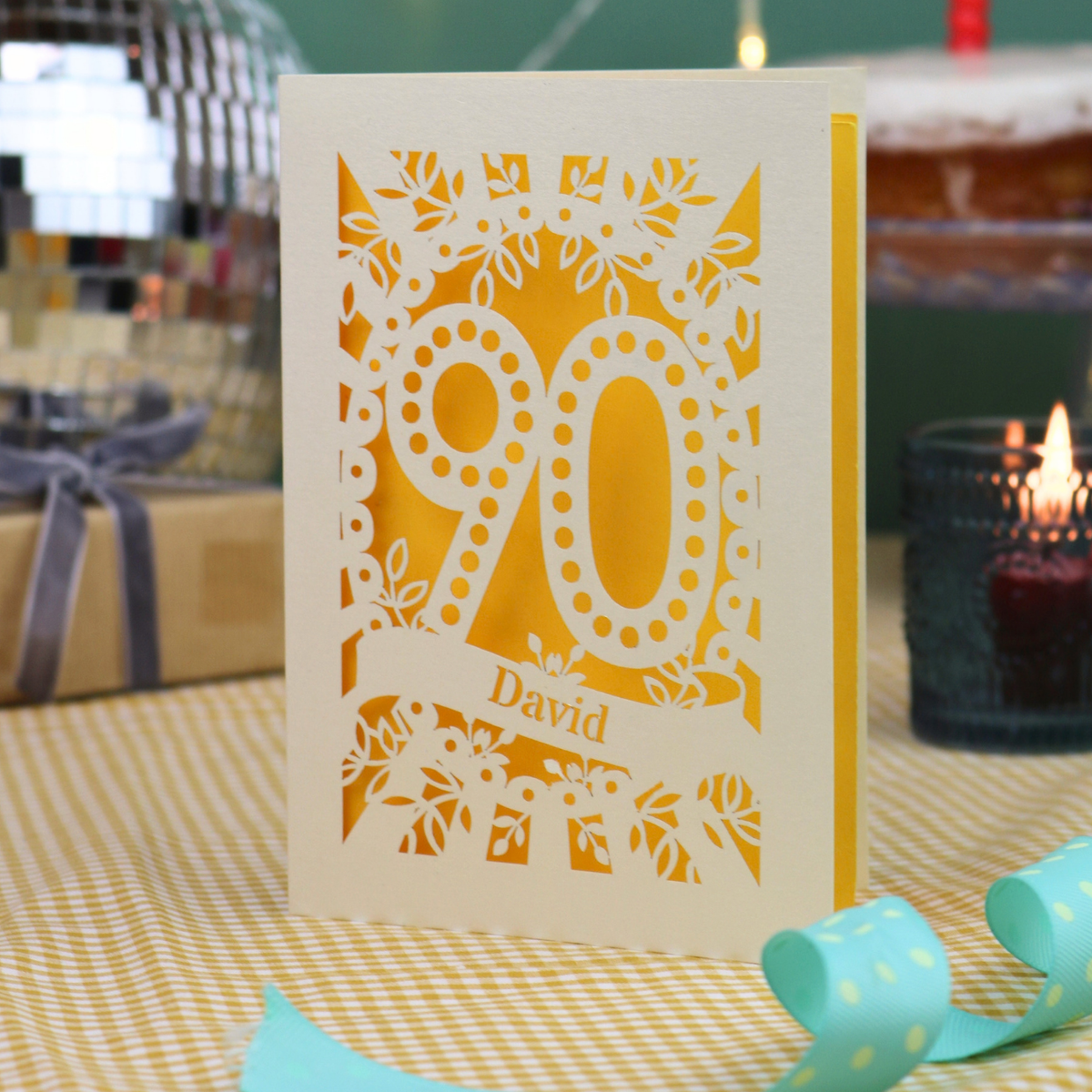 A personalised 90th birthday card with intricate laser cut cream floral and number '90' designs, featuring the name 'David' on a banner. The card is shown standing on a checked tablecloth with a turquoise ribbon, a lit candle, and a disco ball in the background.