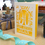 A personalized 90th birthday card with a cream and yellow laser-cut details, standing on a checked tablecloth with party ribbons and a wrapped gift in the background.