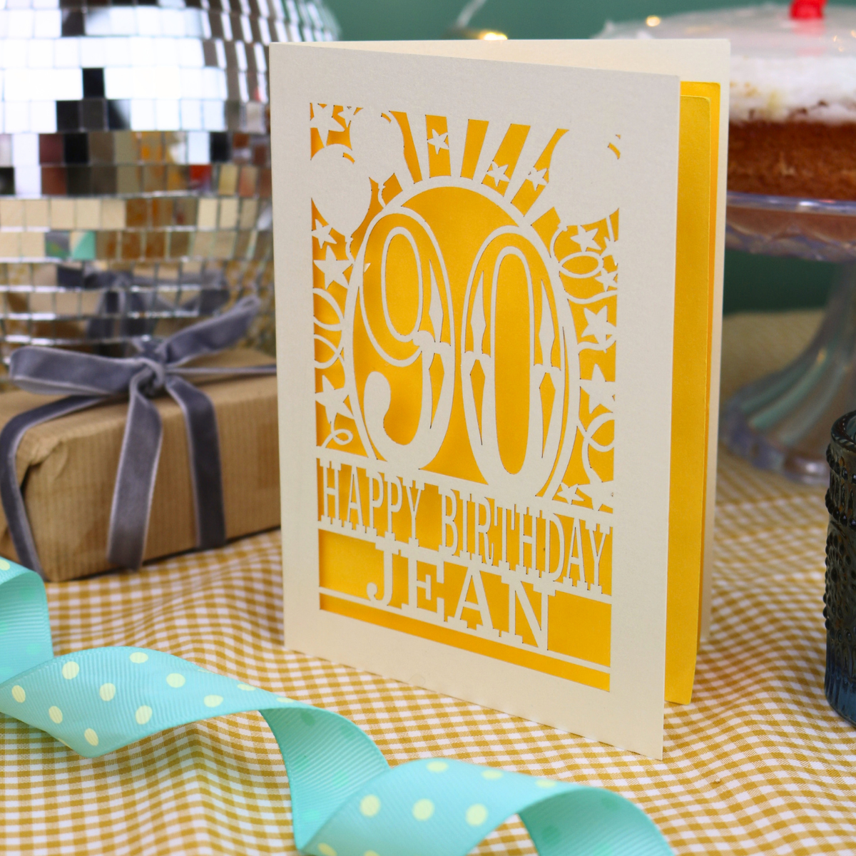 A personalized 90th birthday card with a cream and yellow laser-cut details, standing on a checked tablecloth with party ribbons and a wrapped gift in the background.