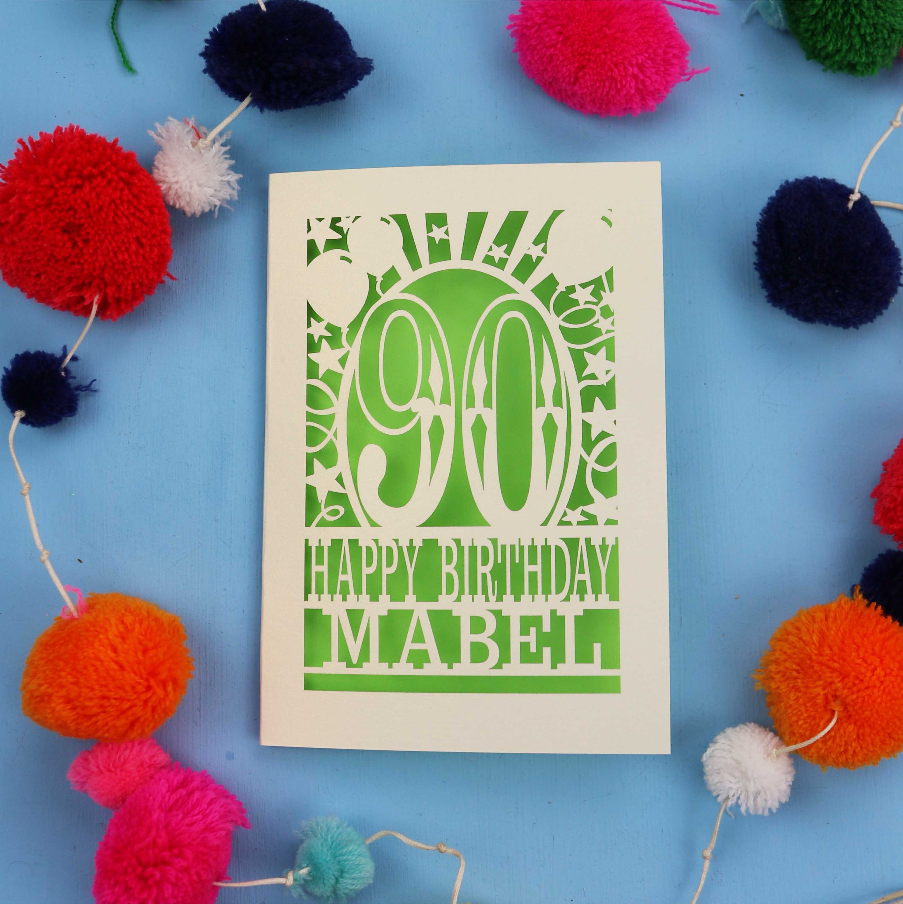 Happy Birthday Mabel - A personalised laser cut 90th birthday card with colourful pom-poms on a blue background