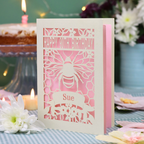 A personalized 90th birthday  card featuring a laser cut bee and honeycomb design with the text 'HAPPY 90TH BIRTHDAY' and 'Sue', displayed on a checked tablecloth with candles and flowers in the background.