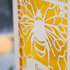 A personalized 90th birthday  card featuring a laser cut bee and honeycomb design with the text 'HAPPY 90TH BIRTHDAY' and 'Sue', displayed on a checked tablecloth with candles and flowers in the background.