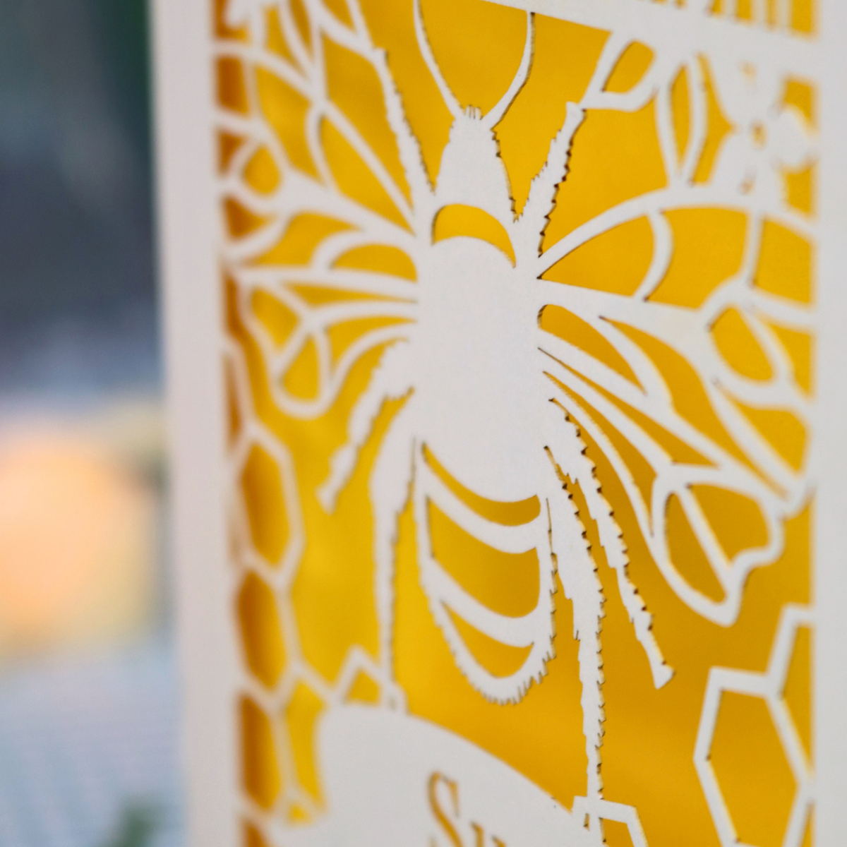 A personalized 90th birthday  card featuring a laser cut bee and honeycomb design with the text 'HAPPY 90TH BIRTHDAY' and 'Sue', displayed on a checked tablecloth with candles and flowers in the background.