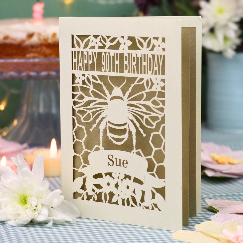 A personalized 90th birthday  card featuring a laser cut bee and honeycomb design with the text 'HAPPY 90TH BIRTHDAY' and 'Sue', displayed on a checked tablecloth with candles and flowers in the background.