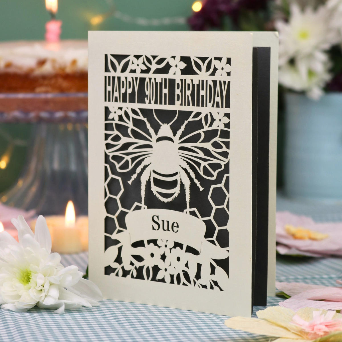 A personalized 90th birthday  card featuring a laser cut bee and honeycomb design with the text 'HAPPY 90TH BIRTHDAY' and 'Sue', displayed on a checked tablecloth with candles and flowers in the background.