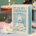 A personalized 90th birthday  card featuring a laser cut bee and honeycomb design with the text 'HAPPY 90TH BIRTHDAY' and 'Sue', displayed on a checked tablecloth with candles and flowers in the background.