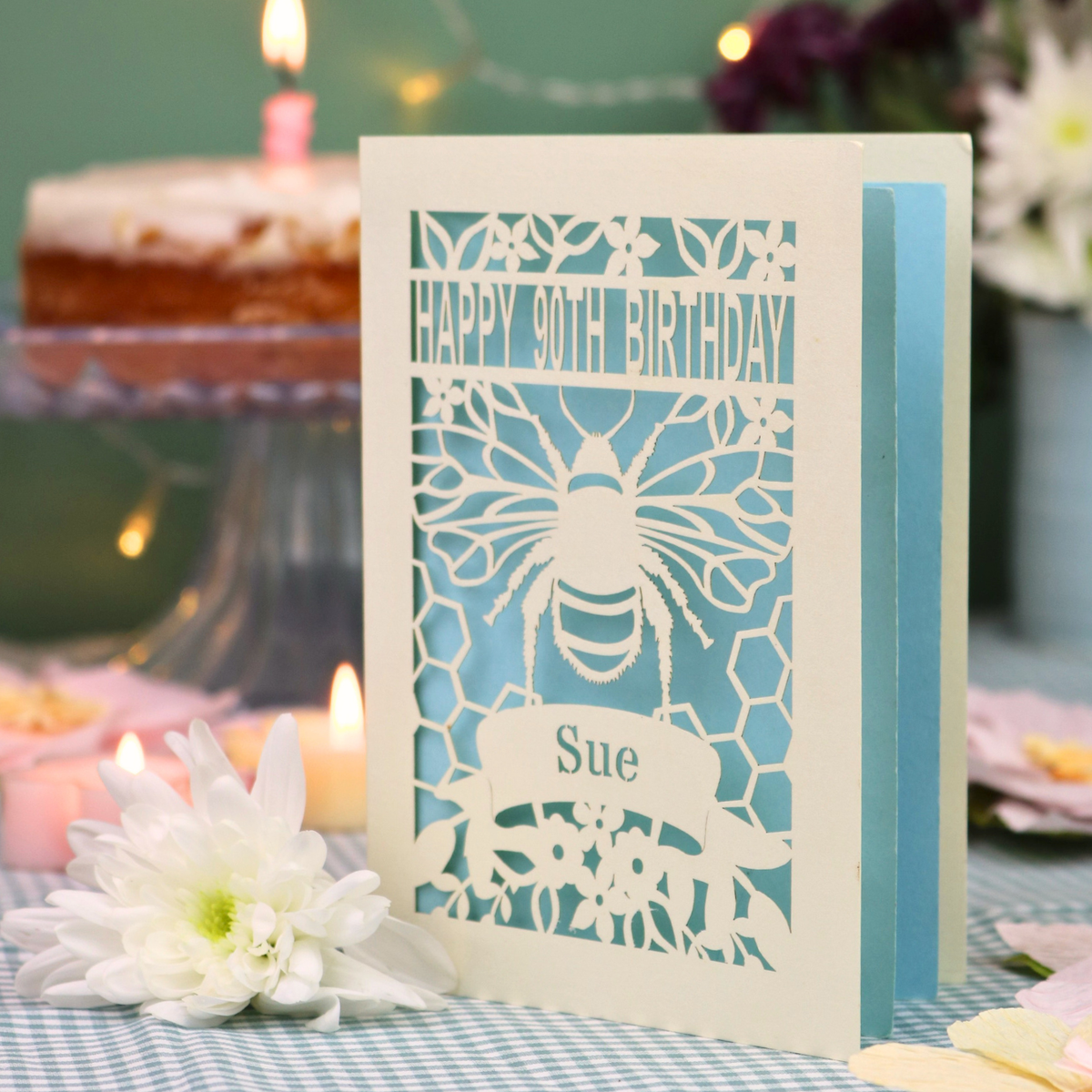 A personalized 90th birthday  card featuring a laser cut bee and honeycomb design with the text 'HAPPY 90TH BIRTHDAY' and 'Sue', displayed on a checked tablecloth with candles and flowers in the background.