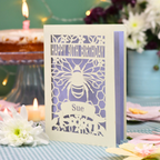 A personalized 90th birthday  card featuring a laser cut bee and honeycomb design with the text 'HAPPY 90TH BIRTHDAY' and 'Sue', displayed on a checked tablecloth with candles and flowers in the background.