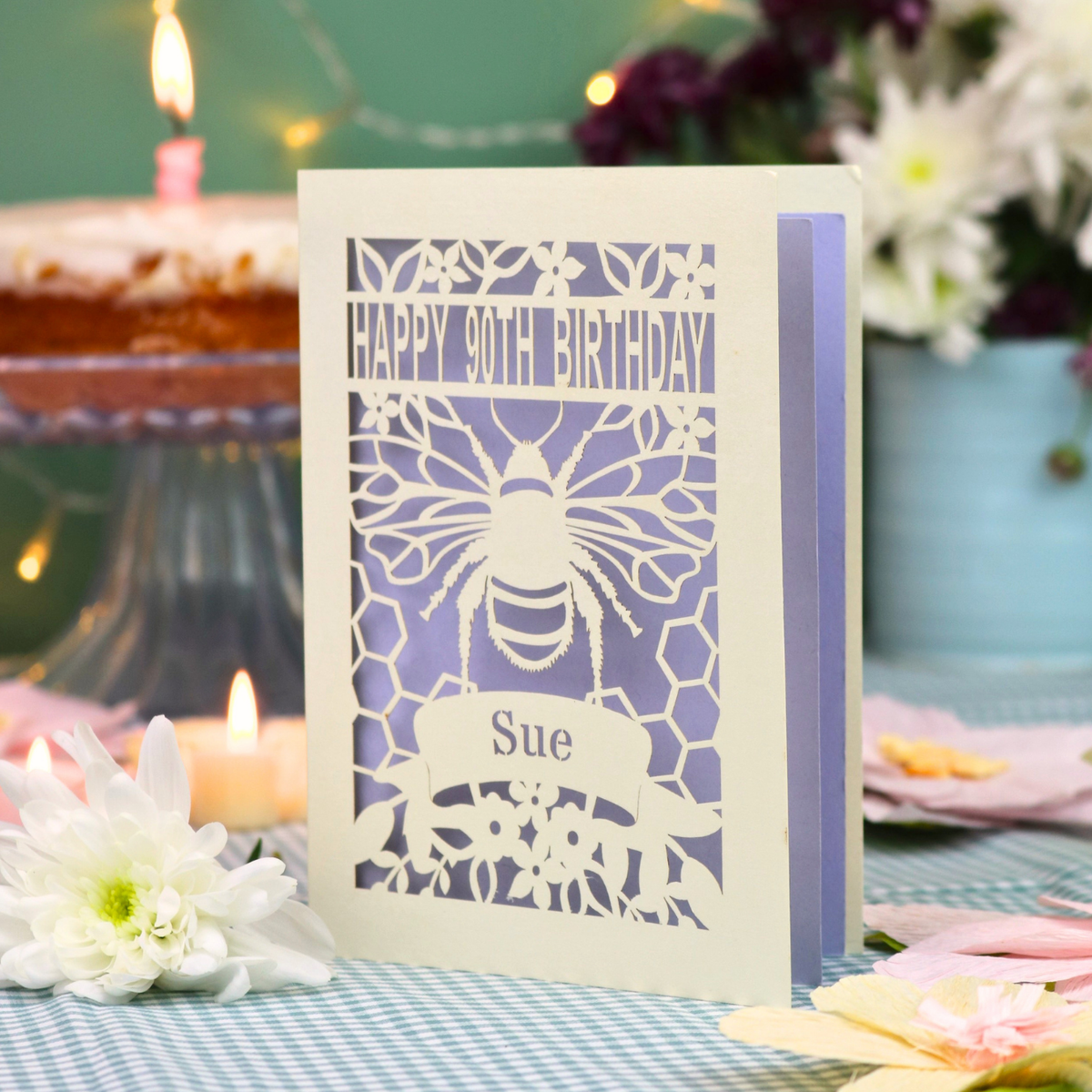 A personalized 90th birthday  card featuring a laser cut bee and honeycomb design with the text 'HAPPY 90TH BIRTHDAY' and 'Sue', displayed on a checked tablecloth with candles and flowers in the background.