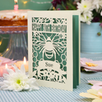 A personalized 90th birthday  card featuring a laser cut bee and honeycomb design with the text 'HAPPY 90TH BIRTHDAY' and 'Sue', displayed on a checked tablecloth with candles and flowers in the background.