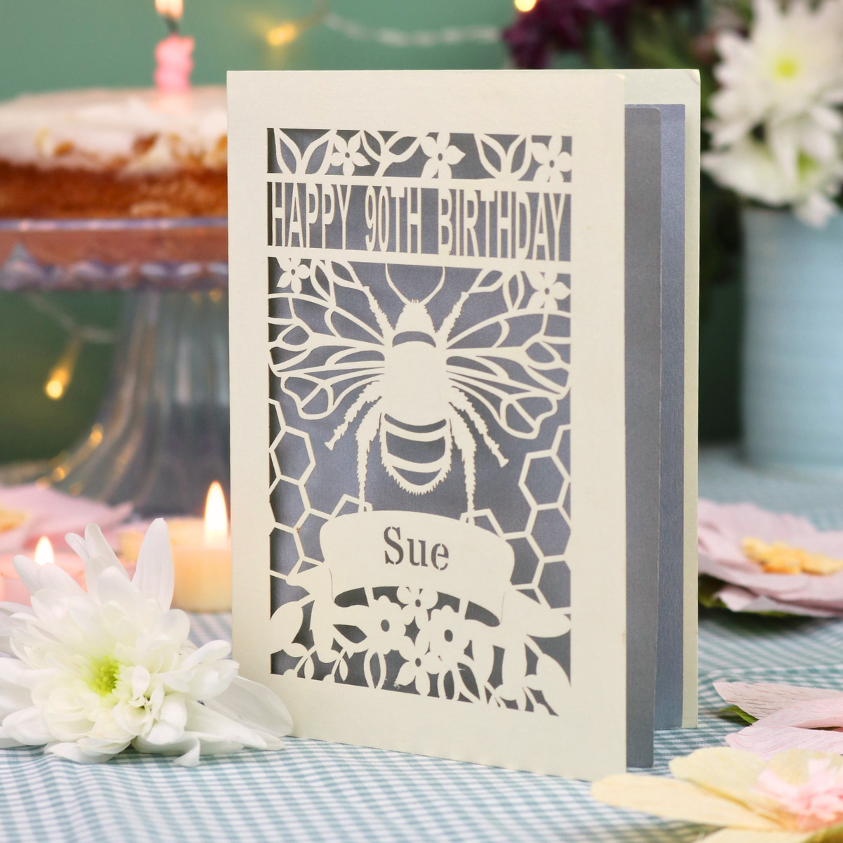 A personalized 90th birthday  card featuring a laser cut bee and honeycomb design with the text 'HAPPY 90TH BIRTHDAY' and 'Sue', displayed on a checked tablecloth with candles and flowers in the background.