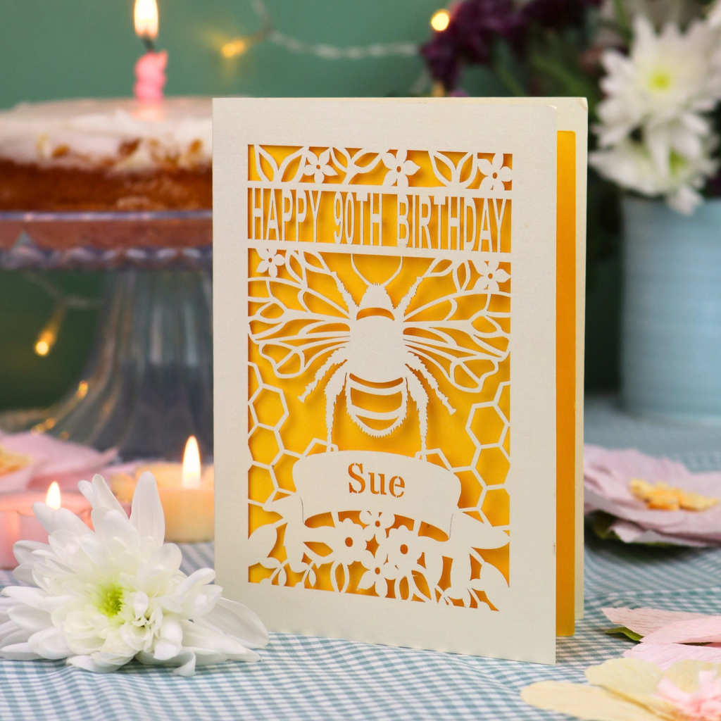 A personalized 90th birthday  card featuring a laser cut bee and honeycomb design with the text 'HAPPY 90TH BIRTHDAY' and 'Sue', displayed on a checked tablecloth with candles and flowers in the background.