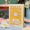 A personalized 90th birthday  card featuring a laser cut bee and honeycomb design with the text 'HAPPY 90TH BIRTHDAY' and 'Sue', displayed on a checked tablecloth with candles and flowers in the background.