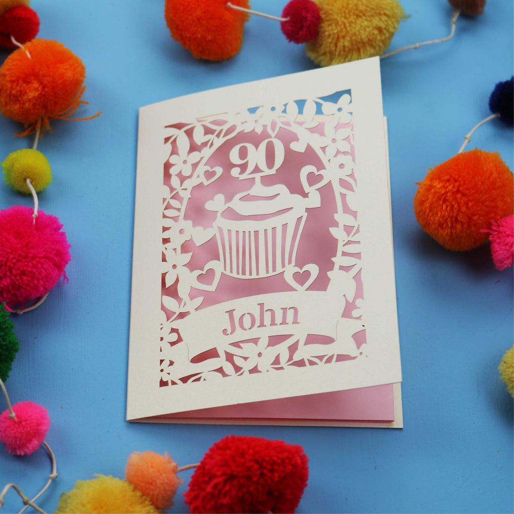 Decorative laser cut 90th birthday card with '90' and 'John' on a blue background with colourful pom-poms.