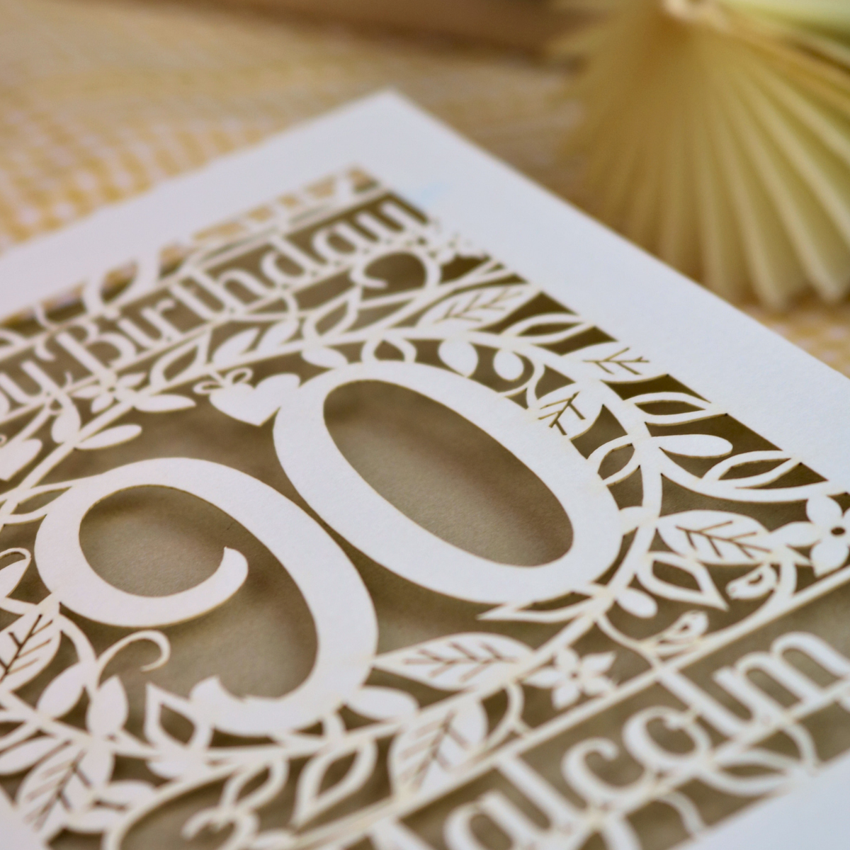 A close-up of a laser-cut birthday card with ornate floral patterns and the number 90, shown on a neutral textured surface.