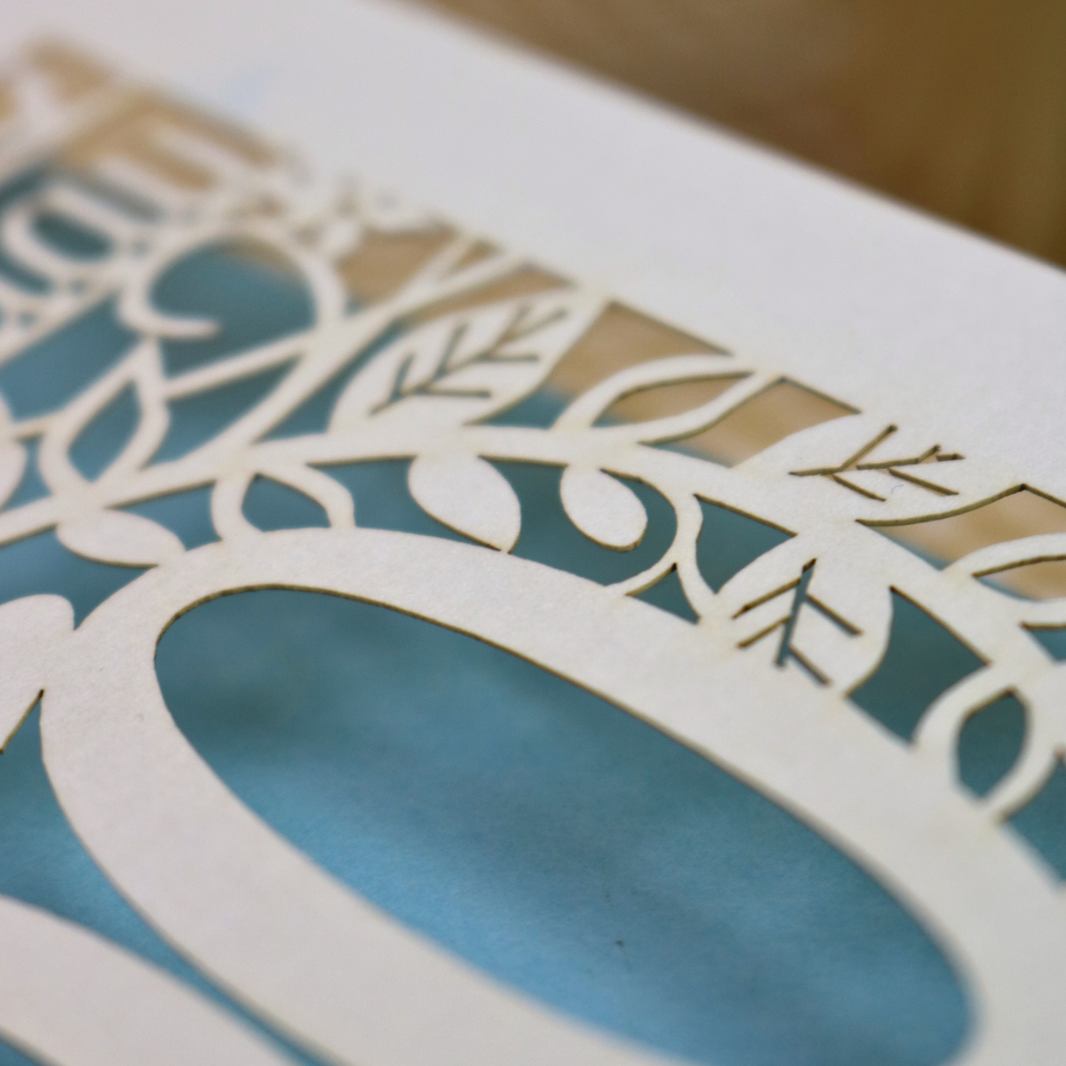 Close-up of a laser-cut birthday card design with intricate leaf and vine patterns, showing a cream cardstock base and blue backing paper visible through the cut outs.