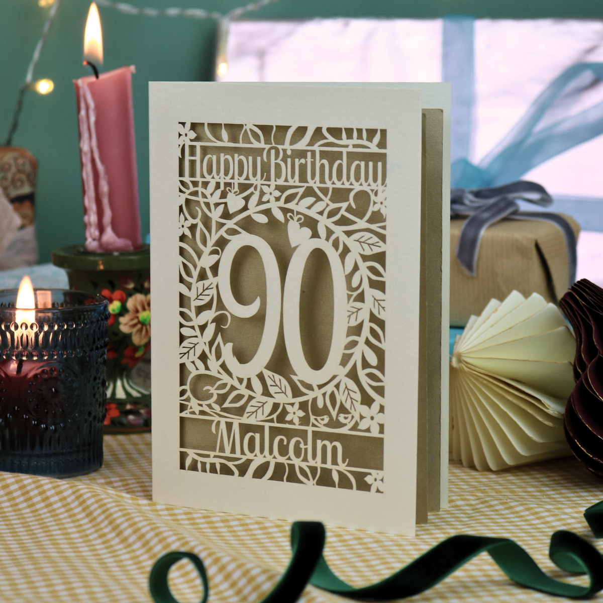 A laser-cut birthday card with 'Happy Birthday 90 Malcolm' displayed on a checked tablecloth with candles, gift boxes, and string lights in the background.
