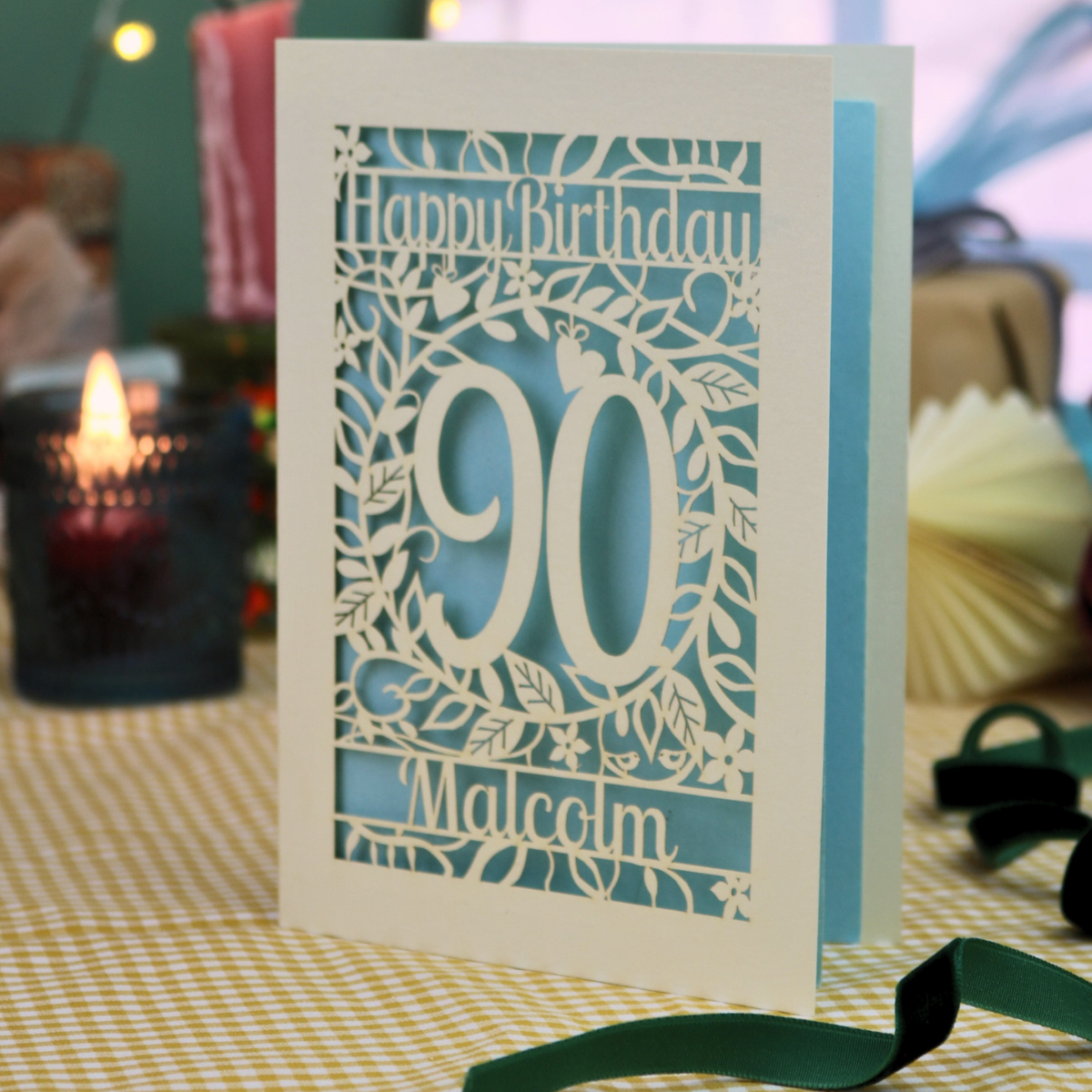 A personalized laser-cut birthday card with a cream border and light blue inner layers, featuring the text 'Happy Birthday 90 Malcolm' surrounded by leaf and vine cut outs. The card is standing on a yellow-and-white checked tablecloth with a lit candle and ribbons in a softly blurred background.