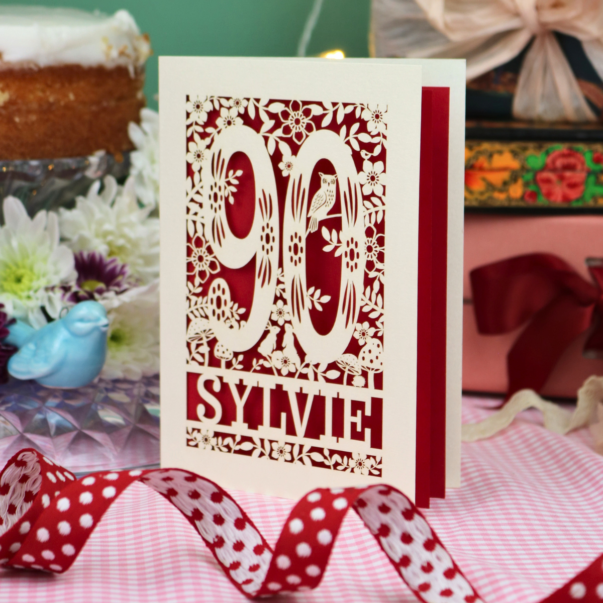 A handmade paper cut 90th birthday card with laser-cut woodland details, the number 90, flowers, and an owl over a dark red background, personalized with the name 'SYLVIE' at the bottom. The card is standing on a pink and white checked tablecloth with a soft-focus background featuring a 90th birthday gifts and a 90th birthday cake.