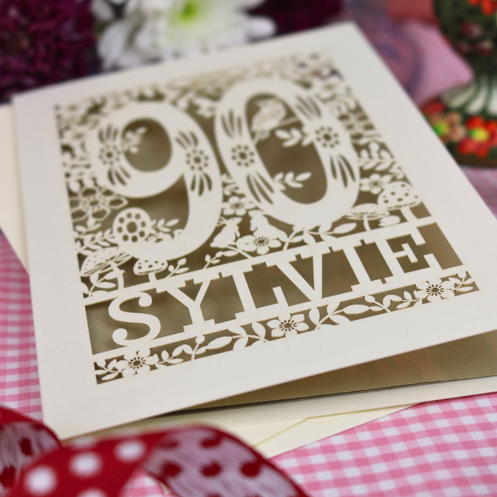 A cut out 90th birthday card with laser-cut woodland details, the number 90, flowers, and an owl over a gold leaf background, personalized with the name 'SYLVIE' at the bottom. 
