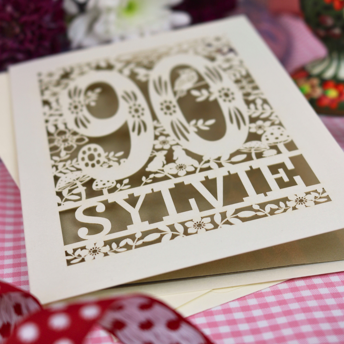 A cut out 90th birthday card with laser-cut woodland details, the number 90, flowers, and an owl over a gold leaf background, personalized with the name 'SYLVIE' at the bottom. 