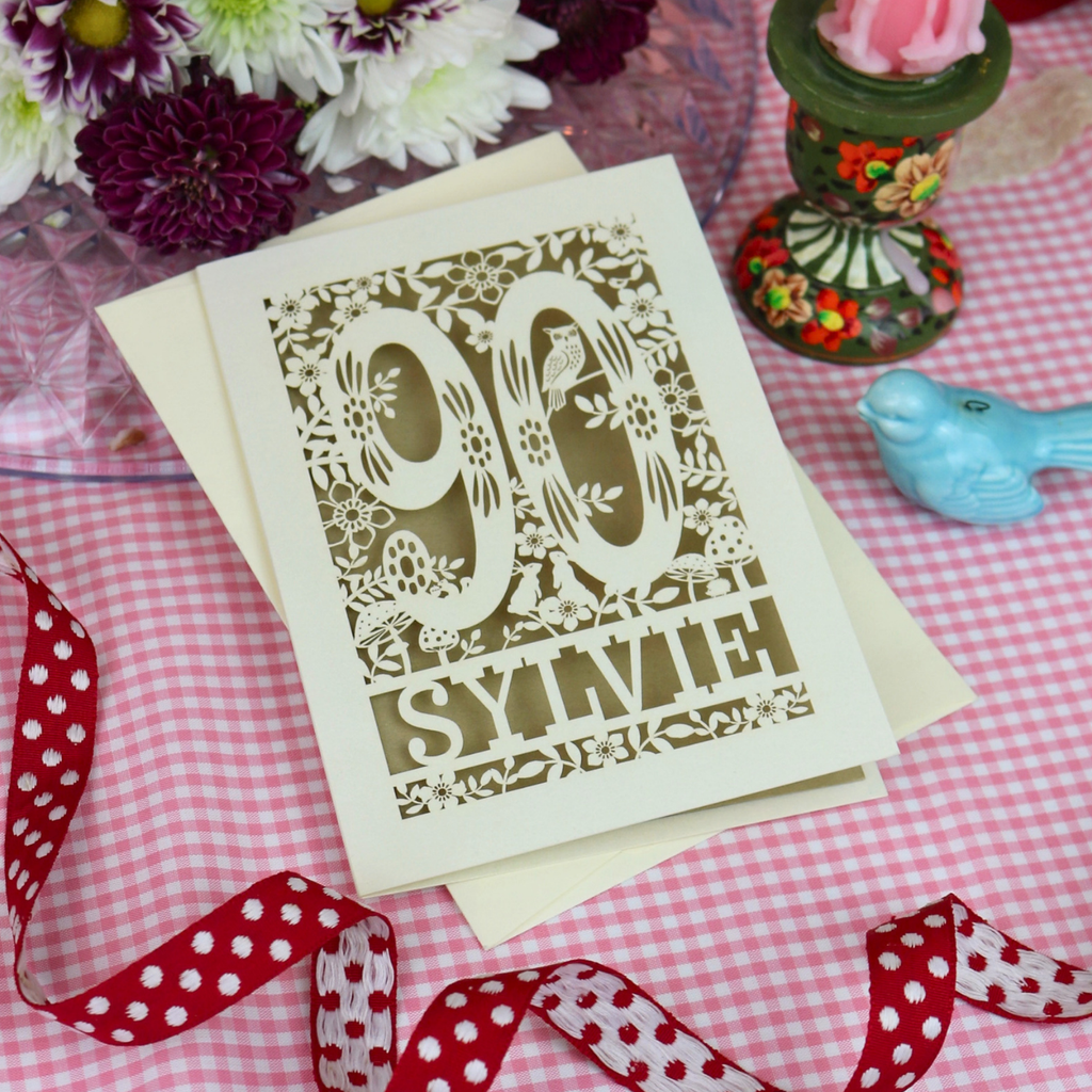 A laser-cut 90th birthday card with floral and bird details, featuring the name 'Sylvie' at the bottom, displayed on a pink and white gingham tablecloth with decorative props including a red polka-dot ribbon, a small blue bird figurine, and flowers.
