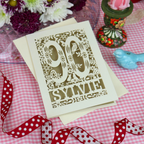 A laser-cut 90th birthday card with floral and bird details, featuring the name 'Sylvie' at the bottom, displayed on a pink and white gingham tablecloth with decorative props including a red polka-dot ribbon, a small blue bird figurine, and flowers.